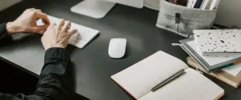 persona tecleando en su computador junto a cuaderno de apuntes